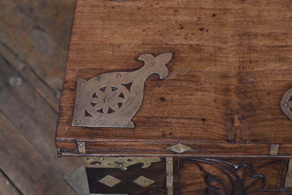 A low rectangular carved wood storage chest featuring decorative brass mounts, corner brackets, studs, and a central relief carving of a floral motif. View IV