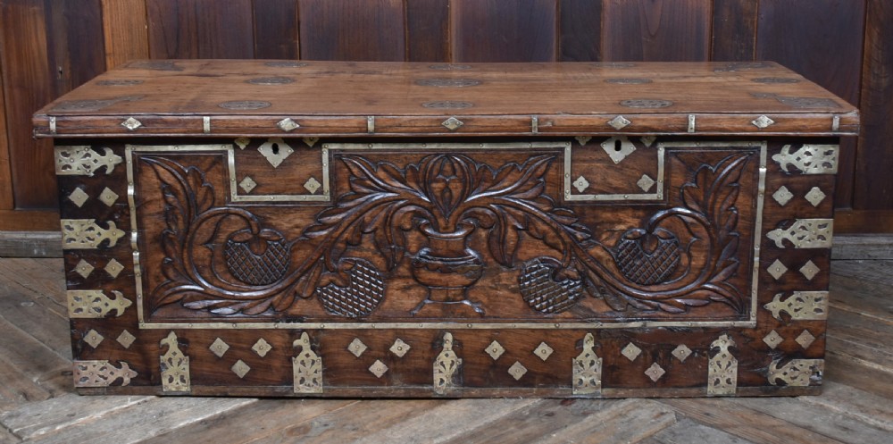 A low rectangular carved wood storage chest featuring decorative brass mounts, corner brackets, studs, and a central relief carving of a floral motif. View VI