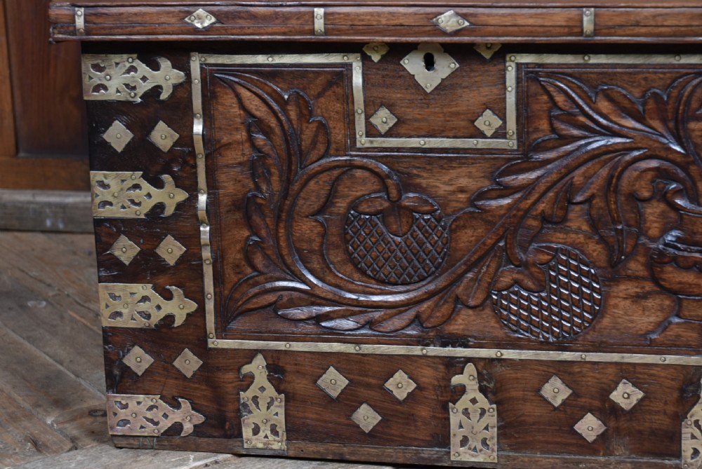 A low rectangular carved wood storage chest featuring decorative brass mounts, corner brackets, studs, and a central relief carving of a floral motif. View VII