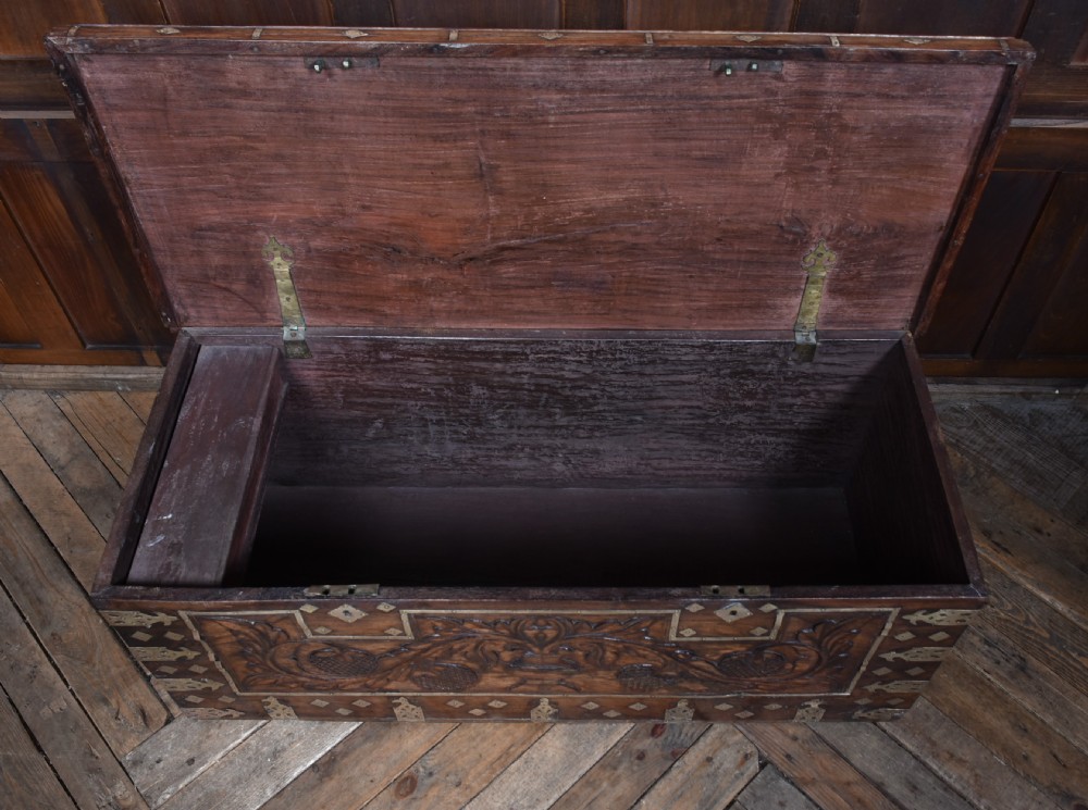 A low rectangular carved wood storage chest featuring decorative brass mounts, corner brackets, studs, and a central relief carving of a floral motif. View X