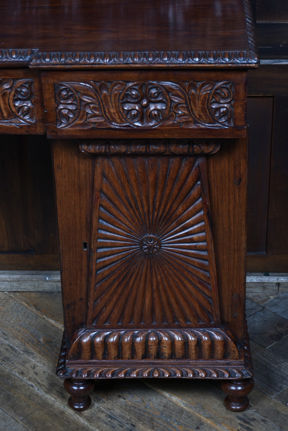 A carved dark padouk wood Anglo-Indian sideboard featuring an ornate foliage gallery top, carved frieze drawers, and two sunburst pedestal doors on turned bun feet. View II
