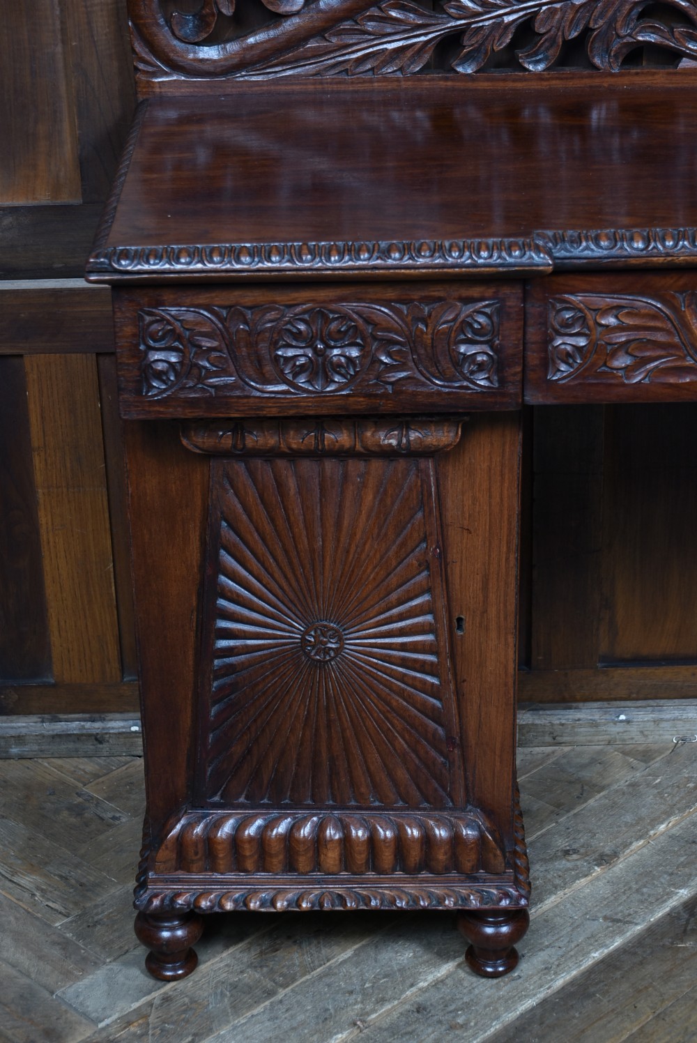 A carved dark padouk wood Anglo-Indian sideboard featuring an ornate foliage gallery top, carved frieze drawers, and two sunburst pedestal doors on turned bun feet. View III