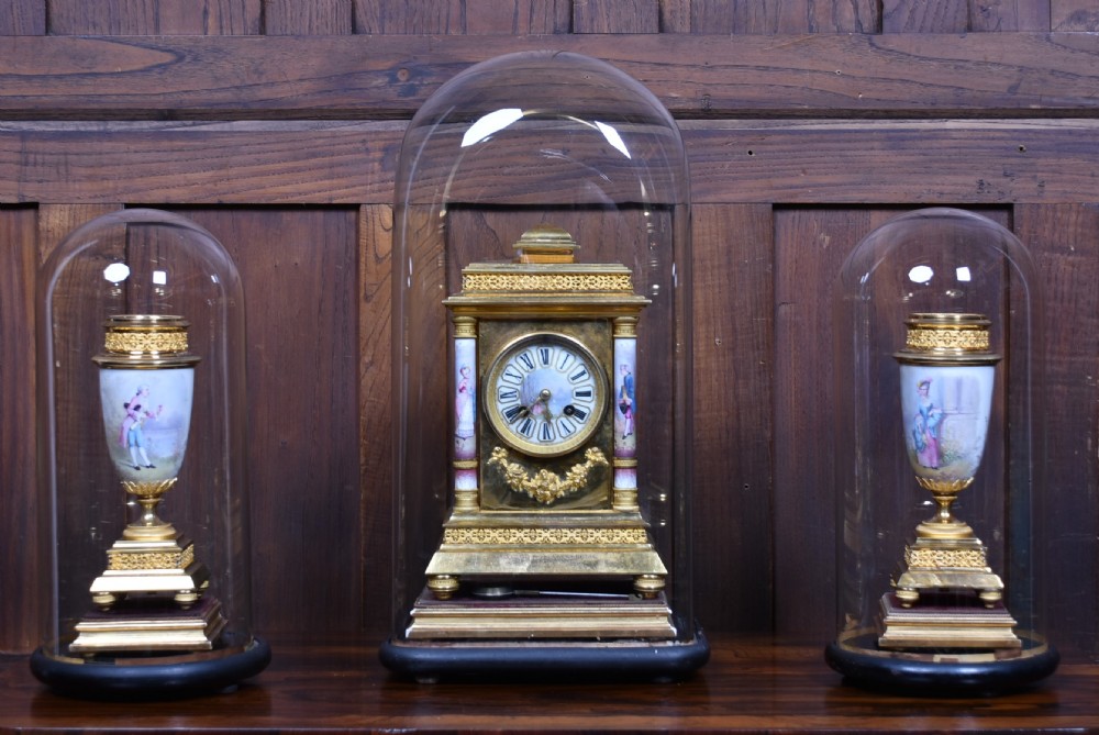 A three-piece antique French gilt metal and painted porcelain clock garniture set with matching side urns, each housed under a glass dome on a black base. View VIII