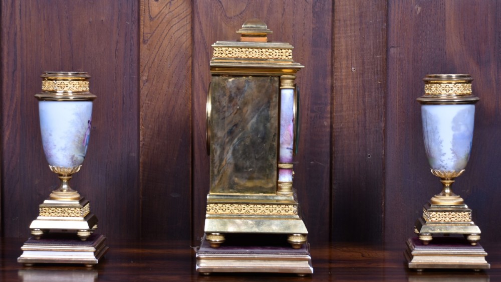 A three-piece antique French gilt metal and painted porcelain clock garniture set with matching side urns, each housed under a glass dome on a black base. View XXII