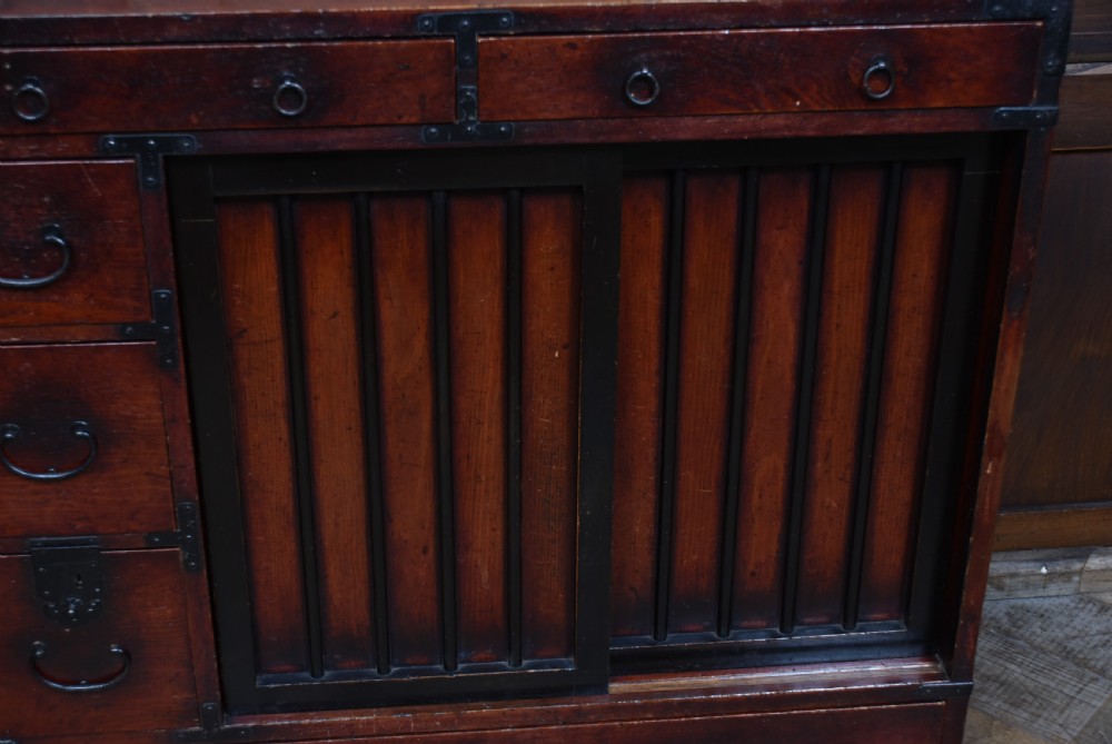 A Japanese Choba Tansu merchant chest featuring dark reddish-brown wood, sliding slatted doors, multiple small drawers, and black iron hardware. View V
