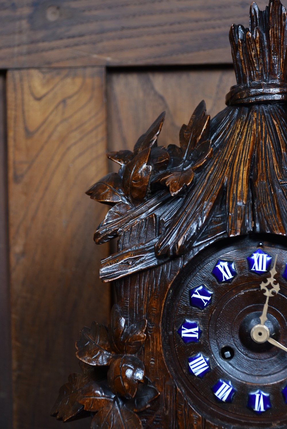 A hand-carved wooden mantel clock featuring a cockerel finial, foliage motifs, and a central dial with blue enamel Roman numeral cartouches. View V