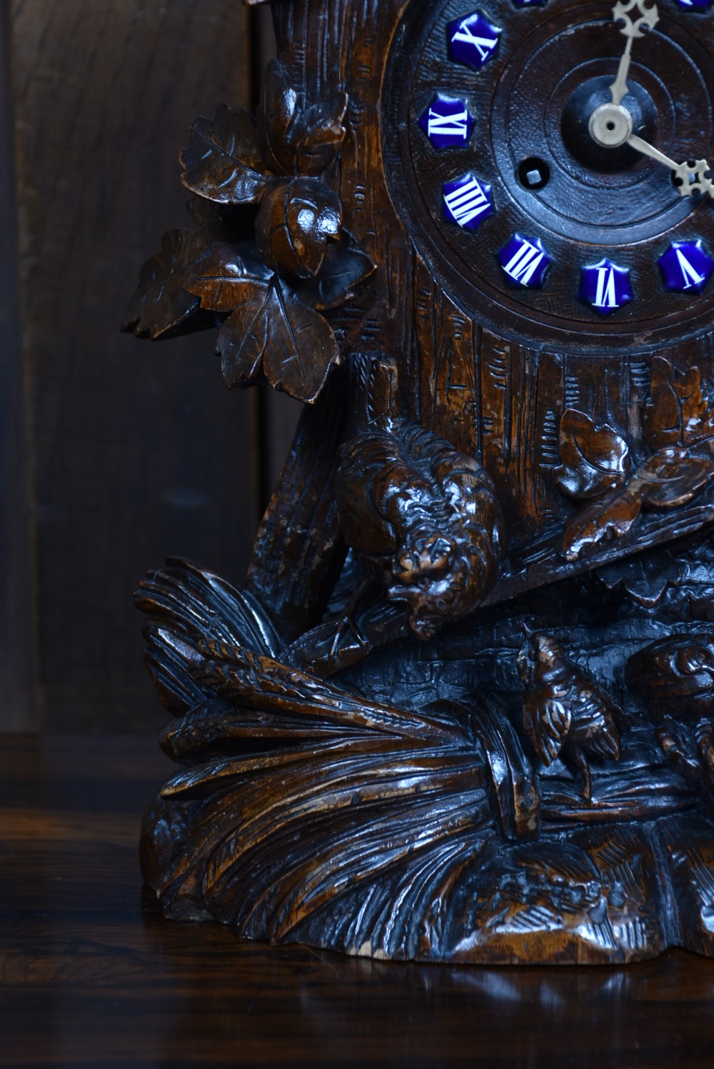 A hand-carved wooden mantel clock featuring a cockerel finial, foliage motifs, and a central dial with blue enamel Roman numeral cartouches. View VI