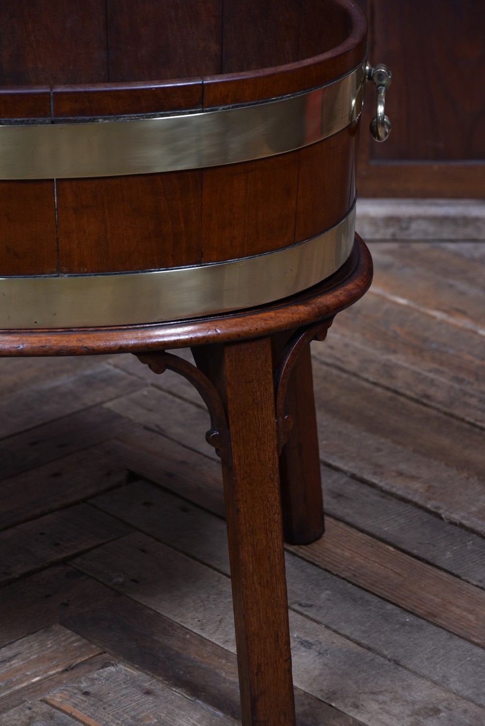 An oval George III mahogany wine cooler bound with two wide brass bands, featuring brass side handles and supported on a fitted wooden stand with four tapered legs. View VI