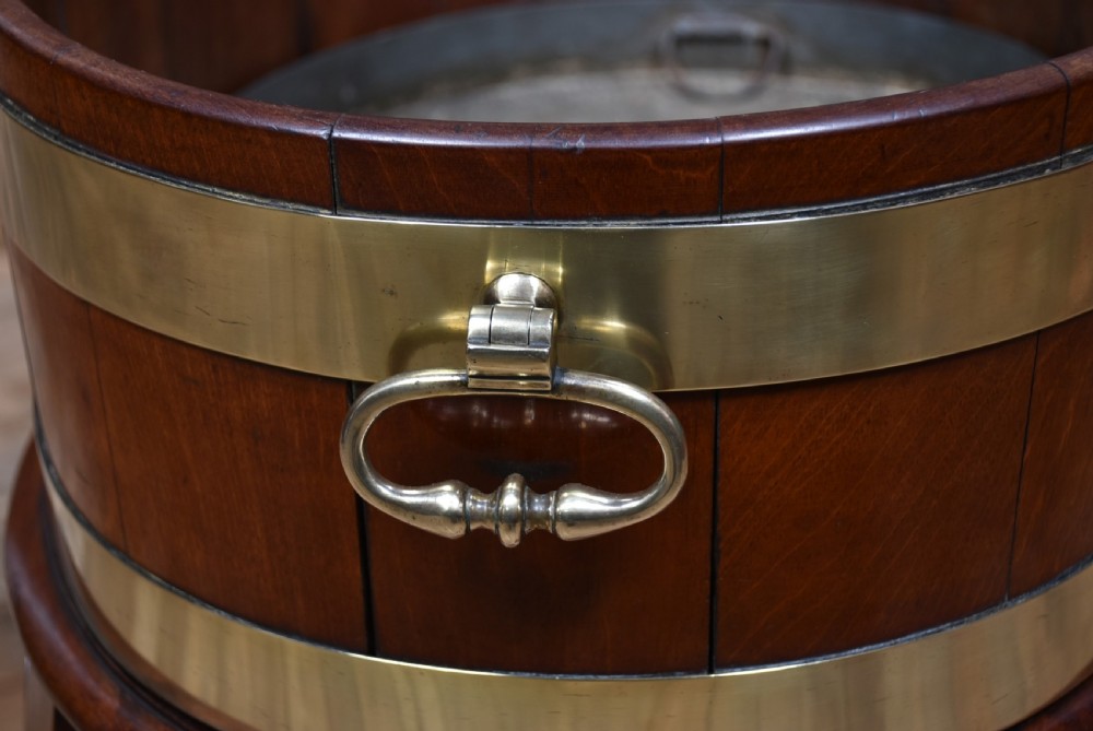 An oval George III mahogany wine cooler bound with two wide brass bands, featuring brass side handles and supported on a fitted wooden stand with four tapered legs. View XI
