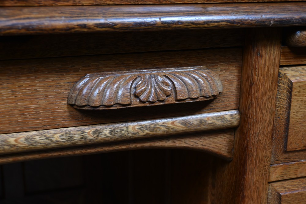 A traditional oak roll-top desk featuring a slatted wooden tambour cover, central kneehole, and twin pedestals fitted with carved handles. View III