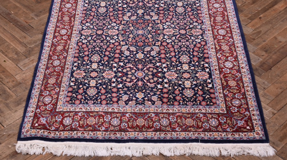 A rectangular hand-knotted wool rug featuring a dark navy central field covered in intricate floral motifs, surrounded by a red border and finished with white fringe edges. View II