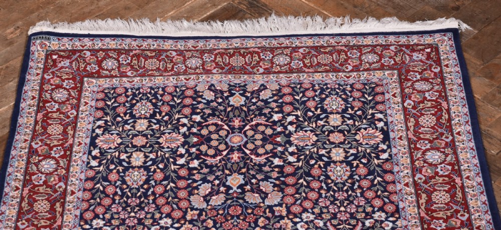 A rectangular hand-knotted wool rug featuring a dark navy central field covered in intricate floral motifs, surrounded by a red border and finished with white fringe edges. View III