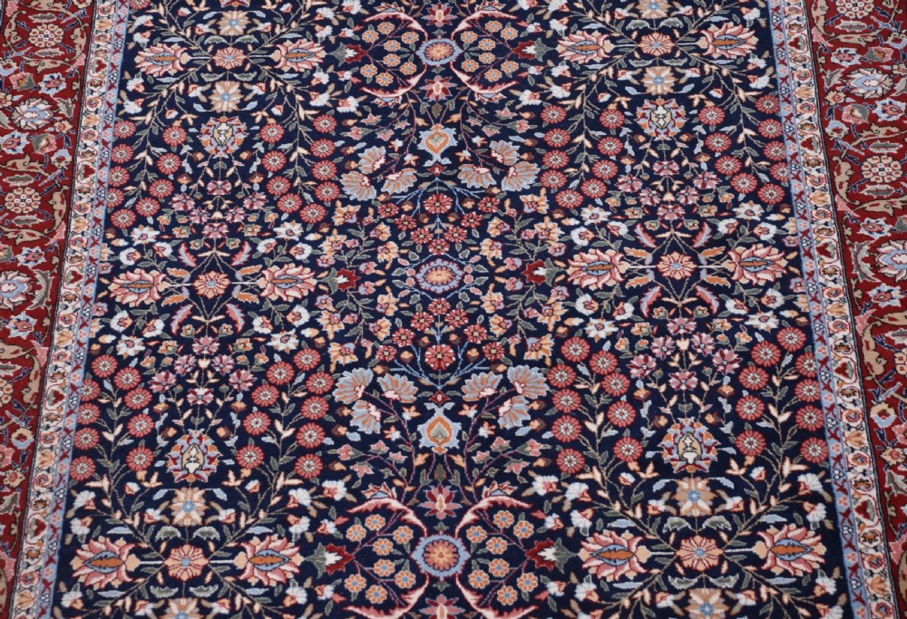 A rectangular hand-knotted wool rug featuring a dark navy central field covered in intricate floral motifs, surrounded by a red border and finished with white fringe edges. View IV