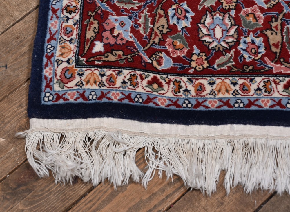 A rectangular hand-knotted wool rug featuring a dark navy central field covered in intricate floral motifs, surrounded by a red border and finished with white fringe edges. View V