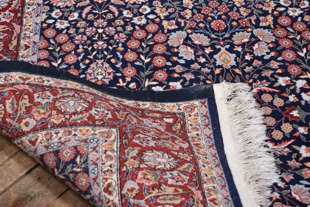 A rectangular hand-knotted wool rug featuring a dark navy central field covered in intricate floral motifs, surrounded by a red border and finished with white fringe edges. View VI