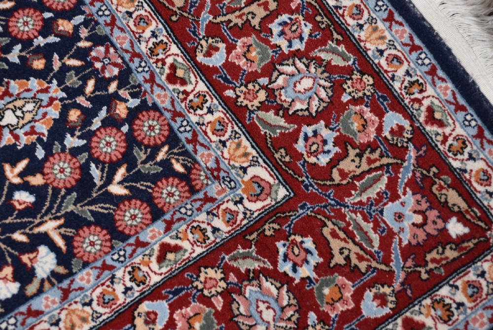 A rectangular hand-knotted wool rug featuring a dark navy central field covered in intricate floral motifs, surrounded by a red border and finished with white fringe edges. View VII