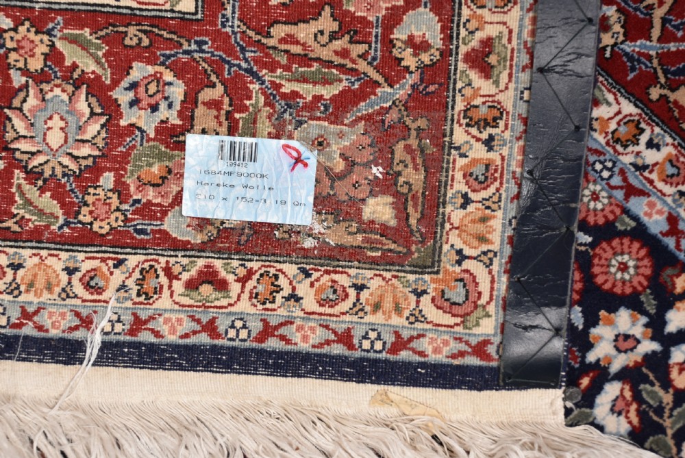 A rectangular hand-knotted wool rug featuring a dark navy central field covered in intricate floral motifs, surrounded by a red border and finished with white fringe edges. View VIII