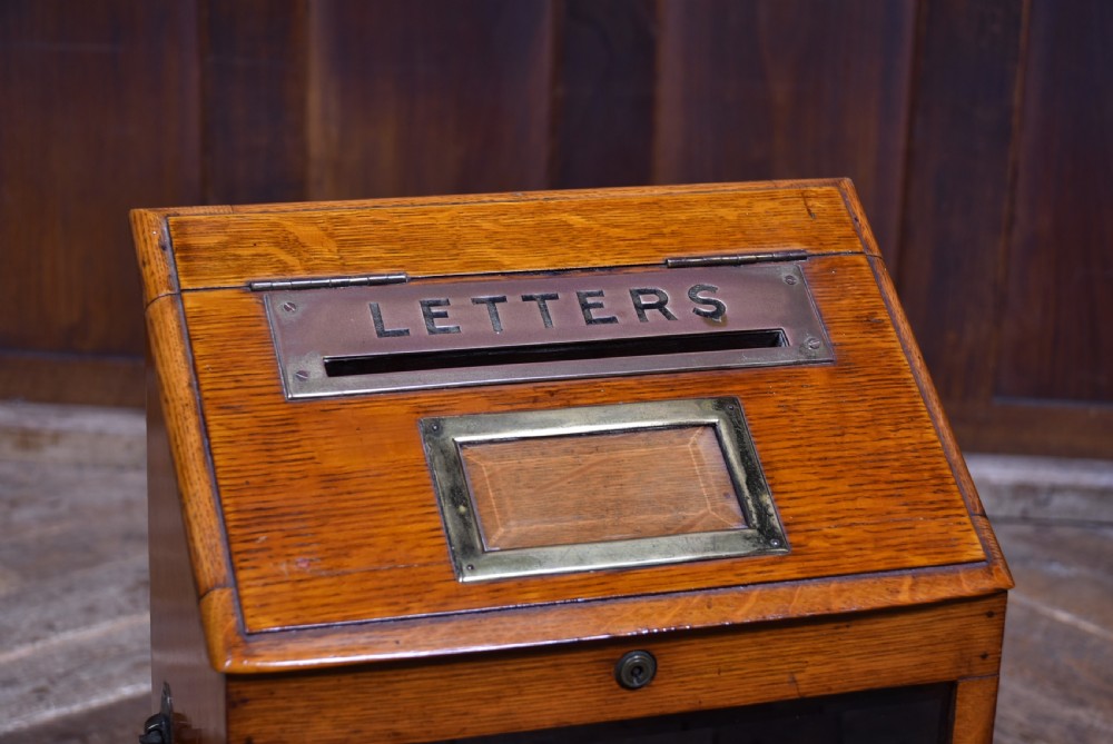 An Edwardian oak desktop letter box featuring a slanted top with a brass letter slot, a framed display aperture, lower vertical compartments, and side handles. View II