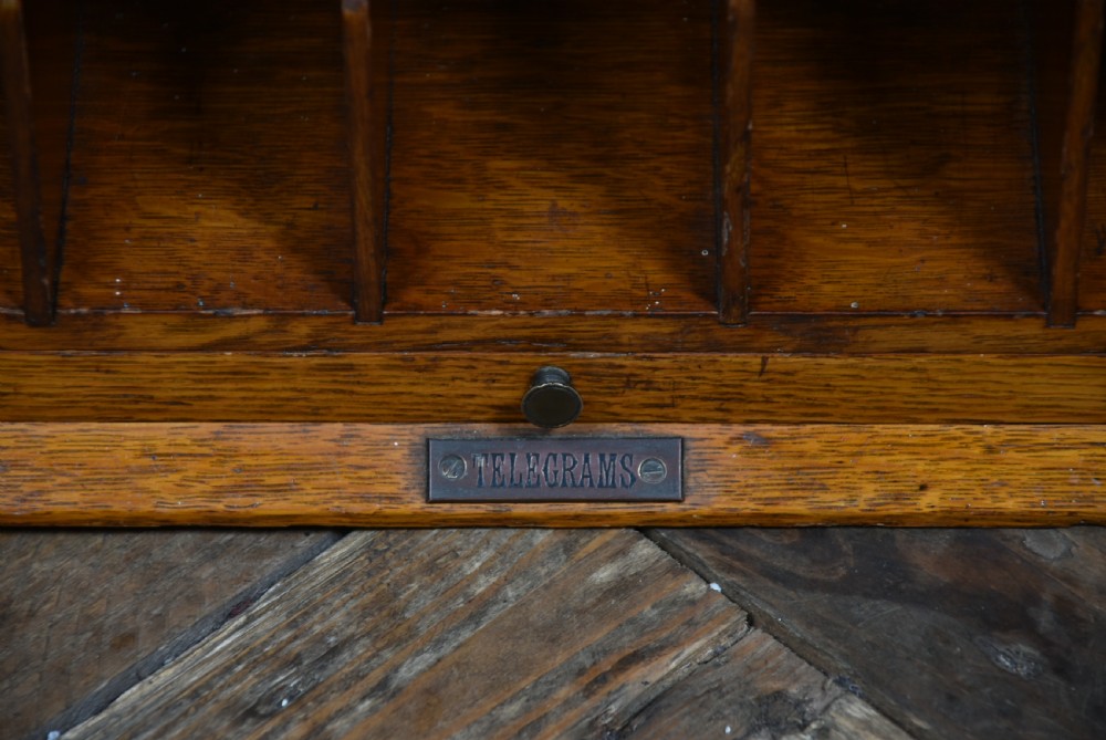 An Edwardian oak desktop letter box featuring a slanted top with a brass letter slot, a framed display aperture, lower vertical compartments, and side handles. View VI