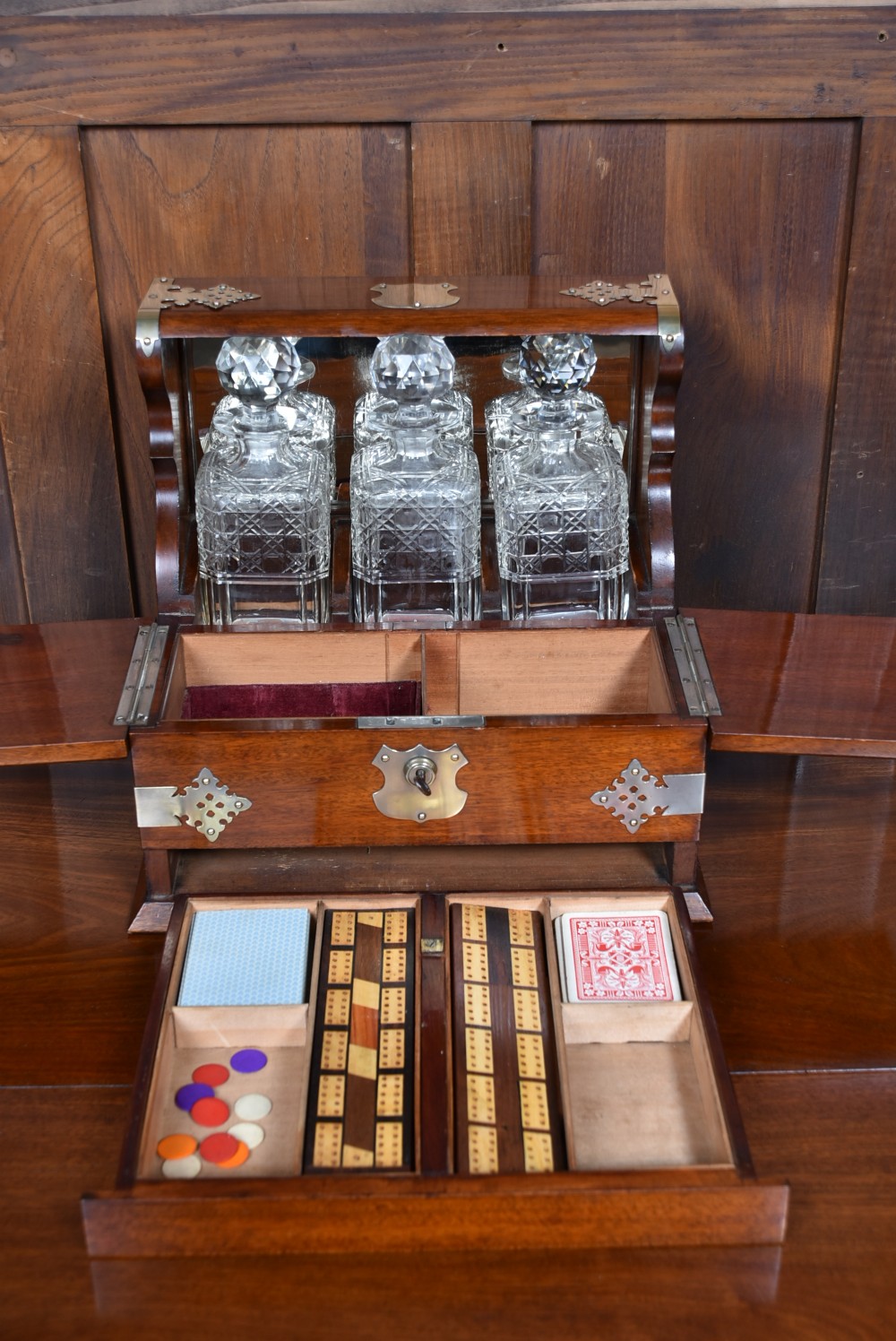 An Edwardian polished mahogany tantalus and games compendium featuring three cut crystal decanters and silver-toned brass strapwork mounts. View XII