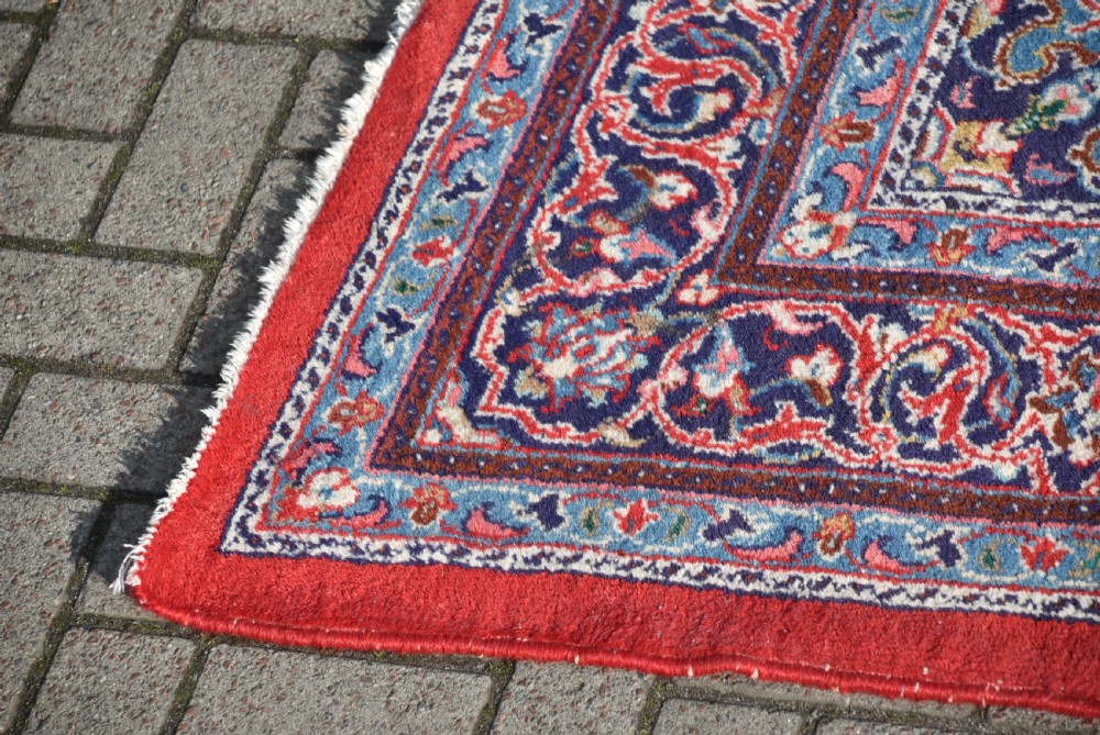 A large hand-knotted vintage Persian rug featuring a central dark blue medallion surrounded by a detailed red field filled with floral arabesque patterns and framed by intricate borders. View V