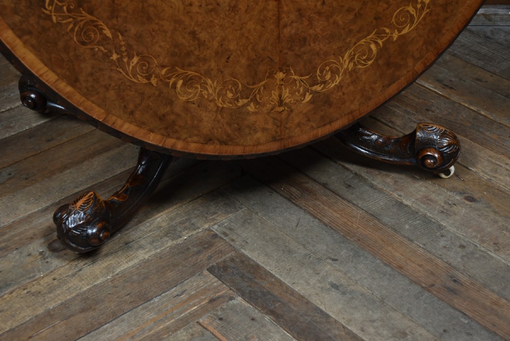 A Victorian oval burr walnut snap-top table featuring detailed floral marquetry inlay and a carved pedestal base displayed upright. View V