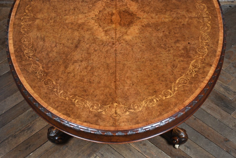 A Victorian oval burr walnut snap-top table featuring detailed floral marquetry inlay and a carved pedestal base displayed upright. View XV
