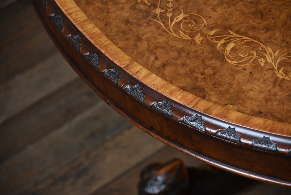 A Victorian oval burr walnut snap-top table featuring detailed floral marquetry inlay and a carved pedestal base displayed upright. View XVI