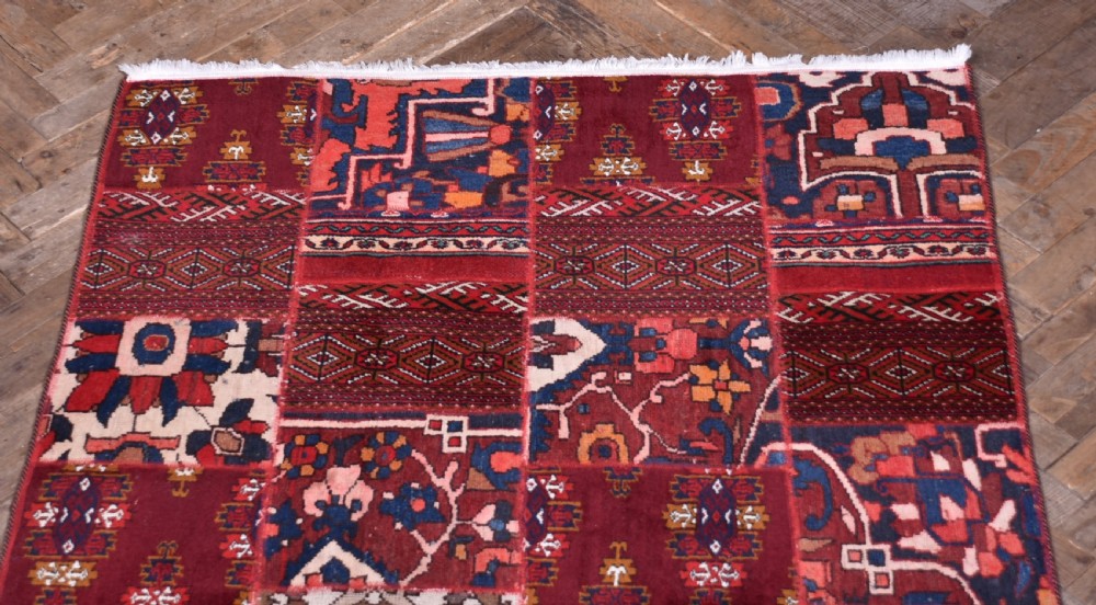 A rectangular hand-stitched Persian patchwork rug featuring geometric and floral panels in deep red, navy blue, and cream with white fringed edges. View IV