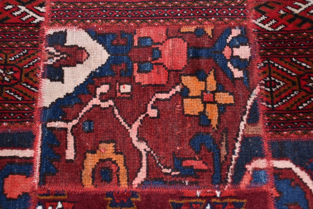 A rectangular hand-stitched Persian patchwork rug featuring geometric and floral panels in deep red, navy blue, and cream with white fringed edges. View VI