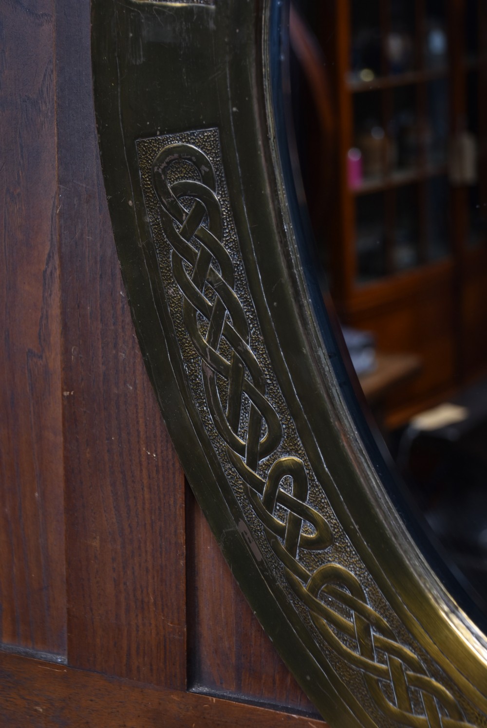 An oval Arts and Crafts brass wall mirror featuring embossed Celtic knotwork interlace detailing around the border frame. View IV