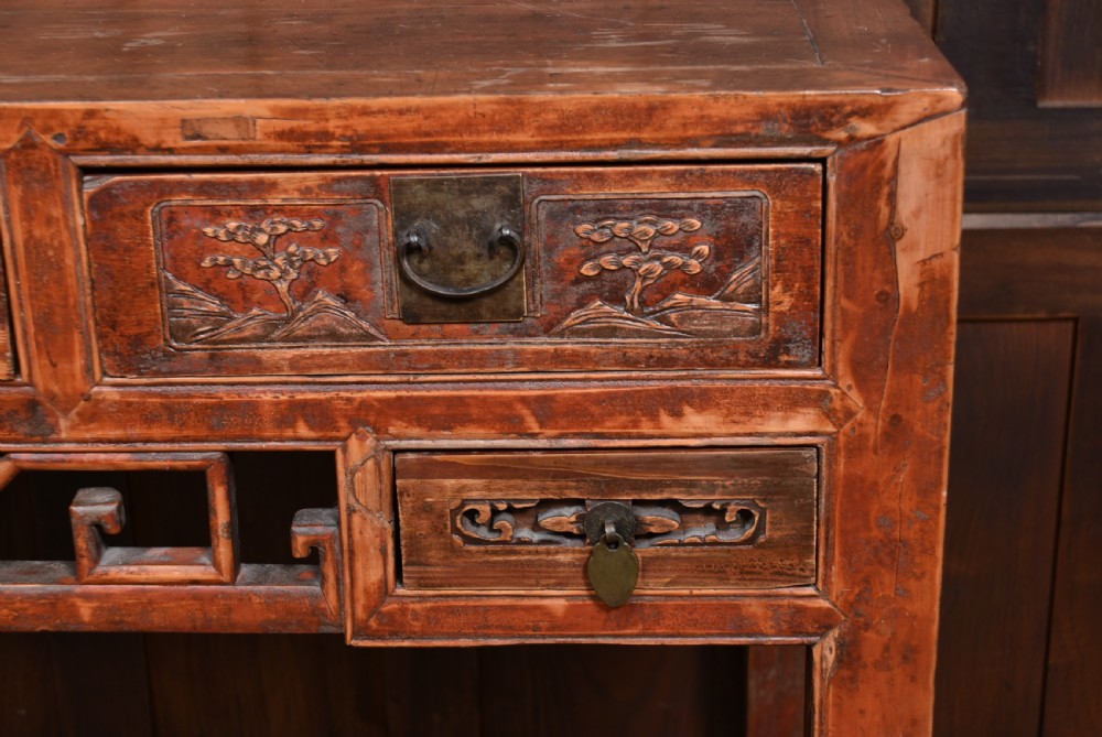 A 19th-century Chinese wooden writing desk featuring carved landscape panels on five drawers, a central openwork fretwork apron, and a warm reddish-brown lacquer finish. View II