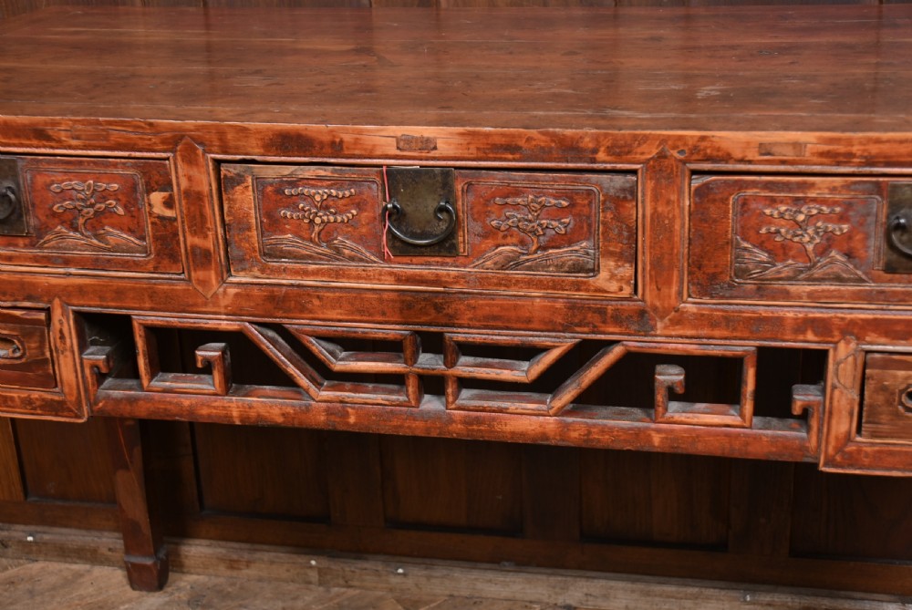 A 19th-century Chinese wooden writing desk featuring carved landscape panels on five drawers, a central openwork fretwork apron, and a warm reddish-brown lacquer finish. View III
