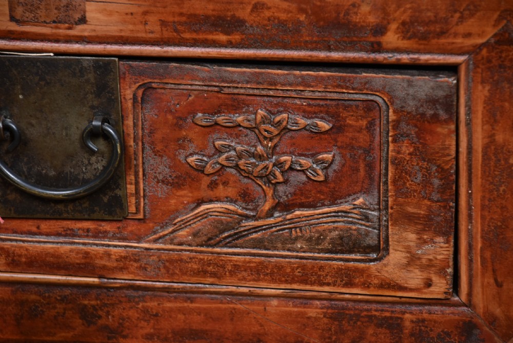 A 19th-century Chinese wooden writing desk featuring carved landscape panels on five drawers, a central openwork fretwork apron, and a warm reddish-brown lacquer finish. View IV