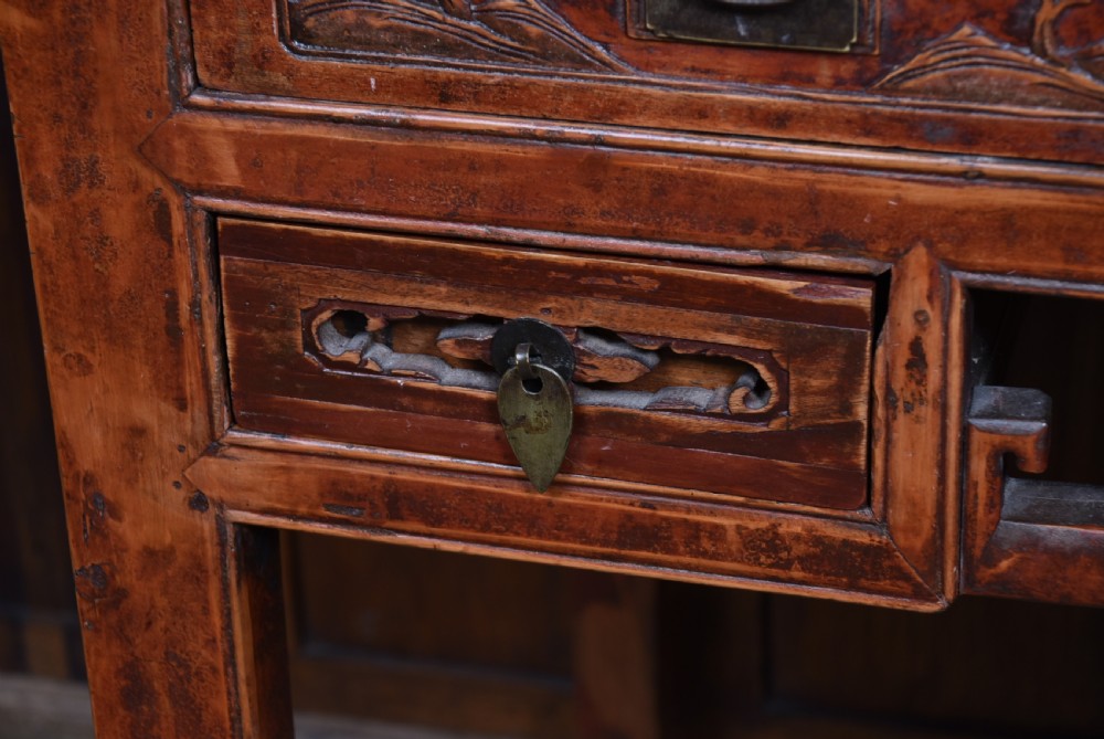 A 19th-century Chinese wooden writing desk featuring carved landscape panels on five drawers, a central openwork fretwork apron, and a warm reddish-brown lacquer finish. View VII