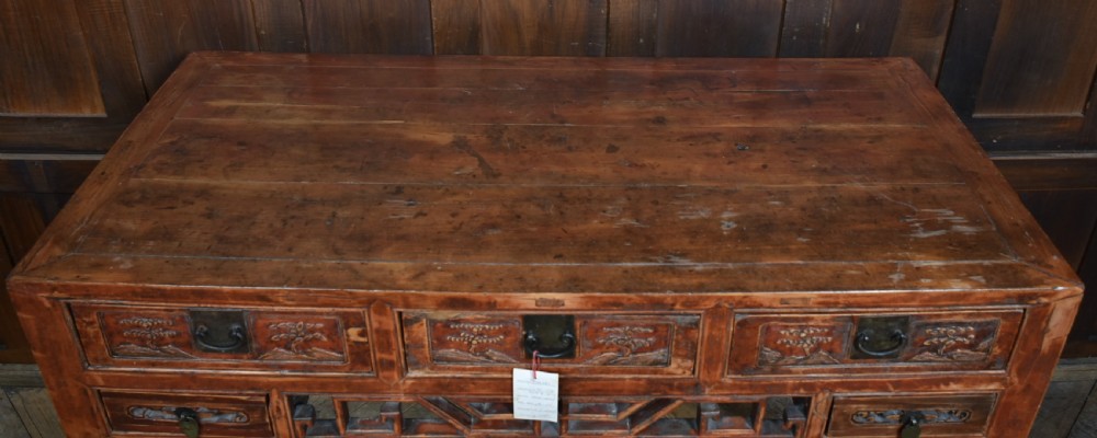 A 19th-century Chinese wooden writing desk featuring carved landscape panels on five drawers, a central openwork fretwork apron, and a warm reddish-brown lacquer finish. View IX