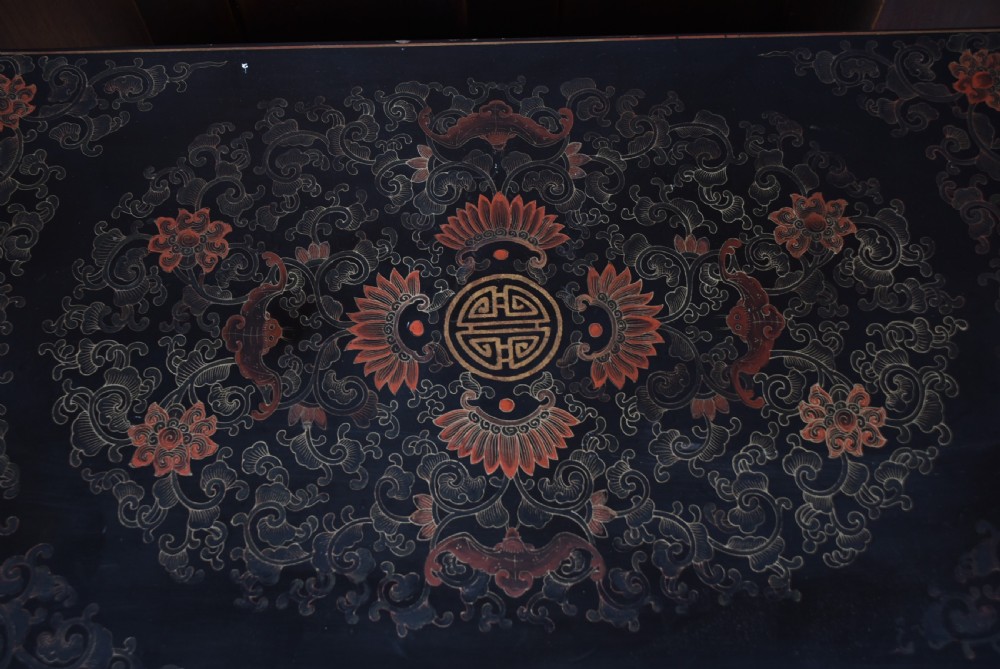 A rectangular black lacquered trunk featuring hand-painted red floral patterns, traditional brass faceplate hardware, and a prominent carved relief panel depicting figures in relief. View IV