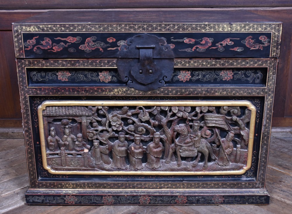 A rectangular black lacquered trunk featuring hand-painted red floral patterns, traditional brass faceplate hardware, and a prominent carved relief panel depicting figures in relief. View V