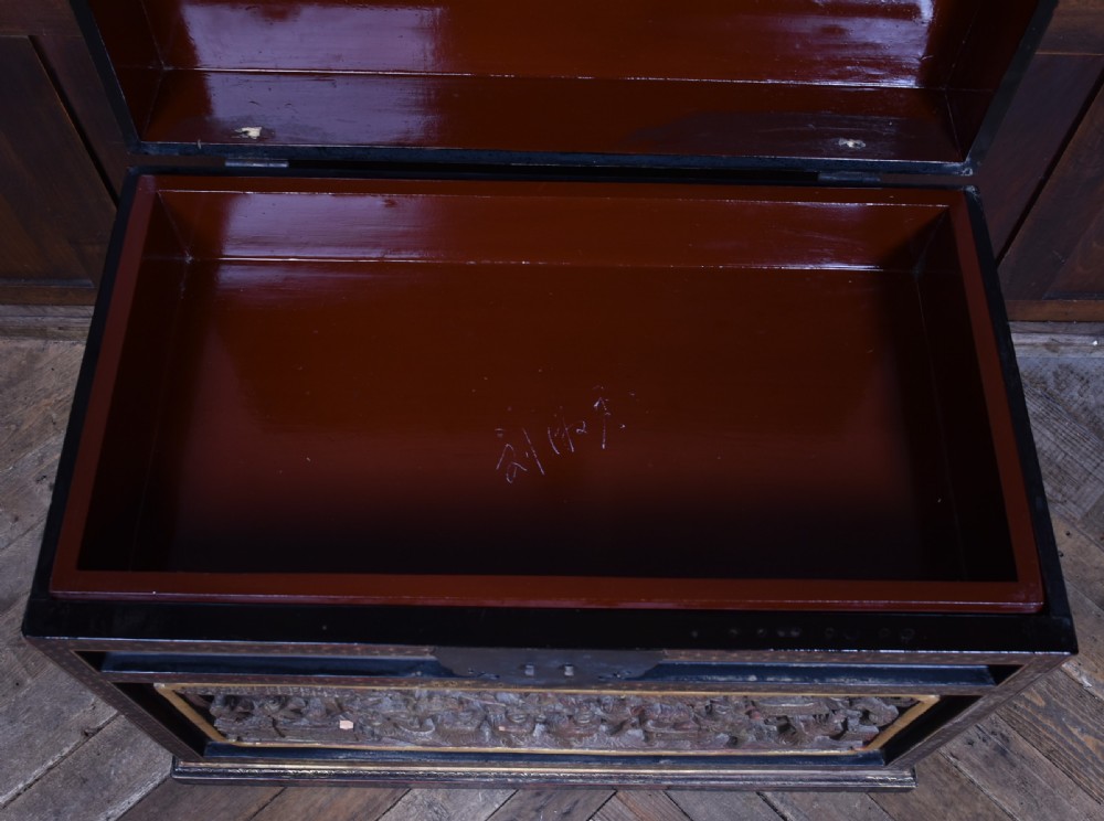 A rectangular black lacquered trunk featuring hand-painted red floral patterns, traditional brass faceplate hardware, and a prominent carved relief panel depicting figures in relief. View VIII