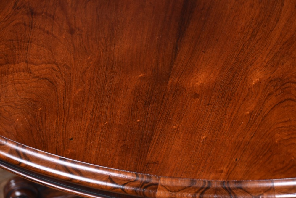 A circular Victorian rosewood tilt-top table featuring a polished figured wood top, carved central pedestal, and ornate scroll legs with castors. View II