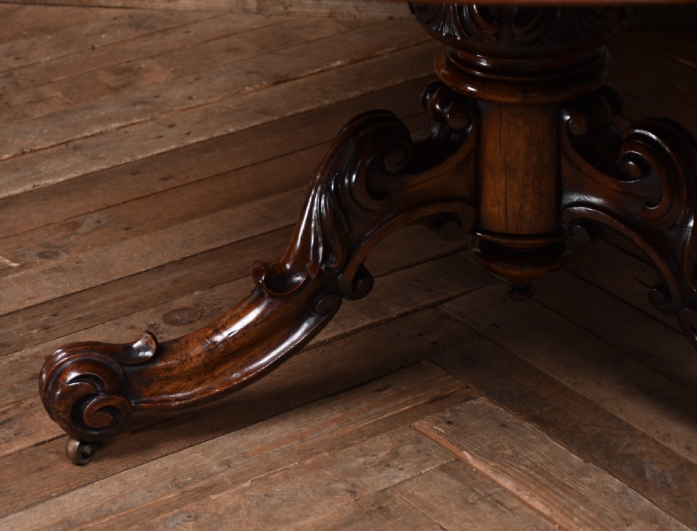 A circular Victorian rosewood tilt-top table featuring a polished figured wood top, carved central pedestal, and ornate scroll legs with castors. View III