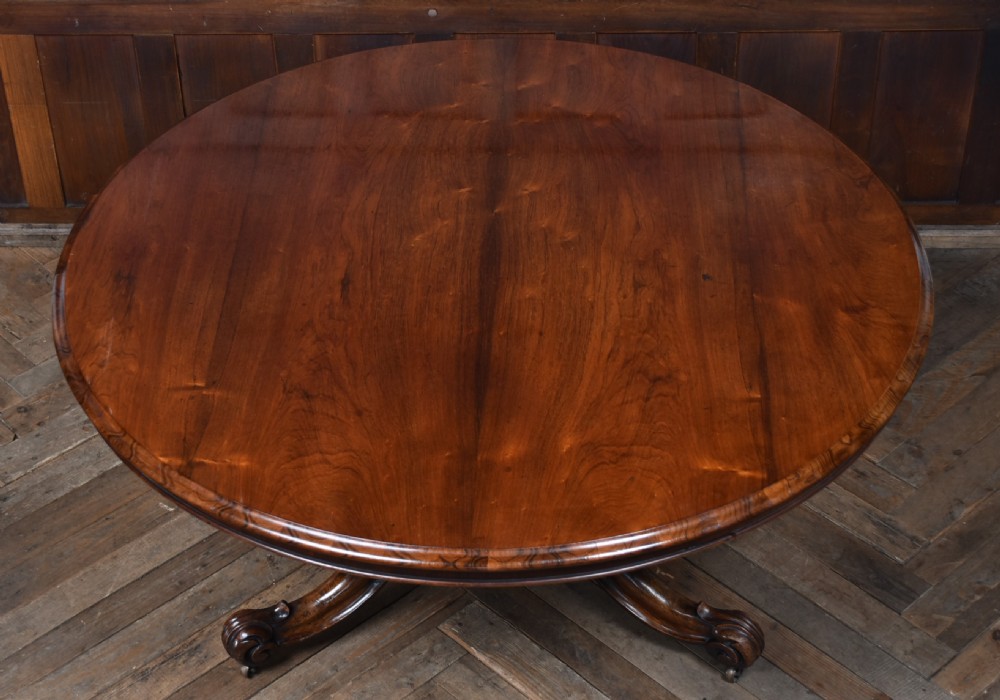 A circular Victorian rosewood tilt-top table featuring a polished figured wood top, carved central pedestal, and ornate scroll legs with castors. View V