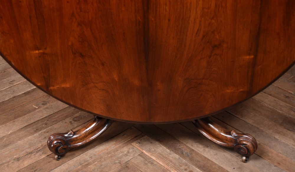 A circular Victorian rosewood tilt-top table featuring a polished figured wood top, carved central pedestal, and ornate scroll legs with castors. View VIII