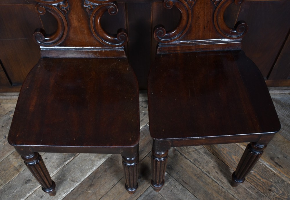 A pair of 19th-century mahogany hall chairs featuring carved scroll backrests, solid wooden seats, and turned fluted front legs. View VII