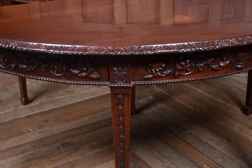 A pair of demilune mahogany console tables featuring carved friezes with swag motifs and four square tapered legs. View IV
