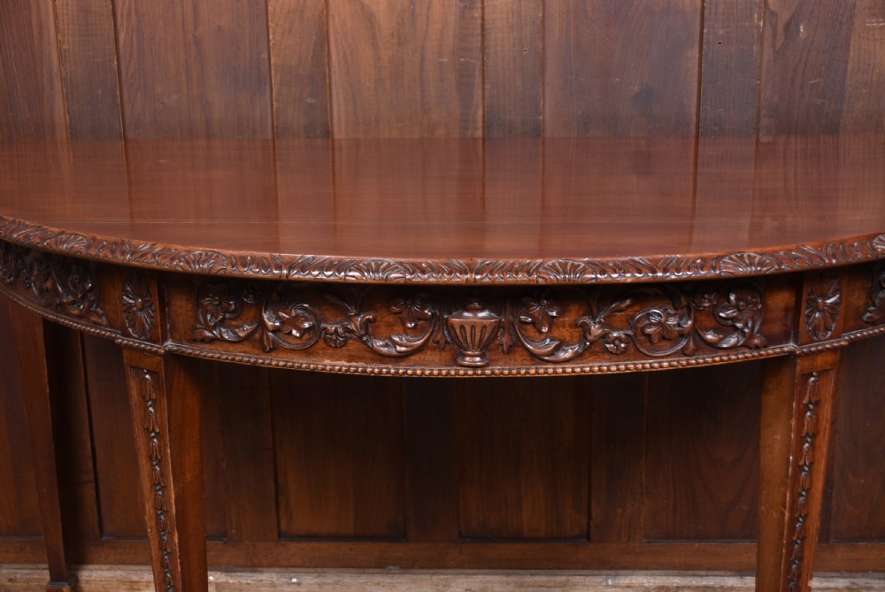 A pair of demilune mahogany console tables featuring carved friezes with swag motifs and four square tapered legs. View VII