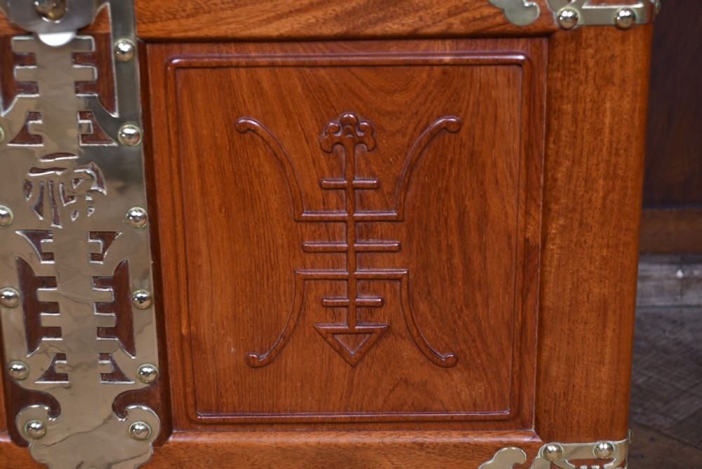A rectangular reddish brown timber blanket box featuring carved Chinese character panels and polished brass corner fittings and straps. View V