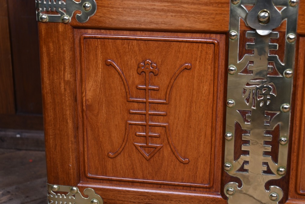 A rectangular reddish brown timber blanket box featuring carved Chinese character panels and polished brass corner fittings and straps. View IX