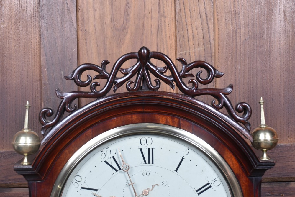 An antique Scottish flame mahogany longcase clock featuring a carved swan-neck pediment with brass finials, a rounded dial, and a veneered trunk door. View VIII