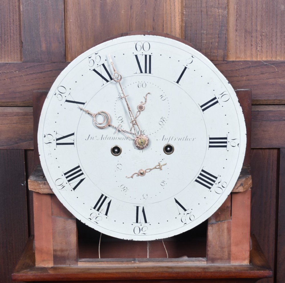 An antique Scottish flame mahogany longcase clock featuring a carved swan-neck pediment with brass finials, a rounded dial, and a veneered trunk door. View IX
